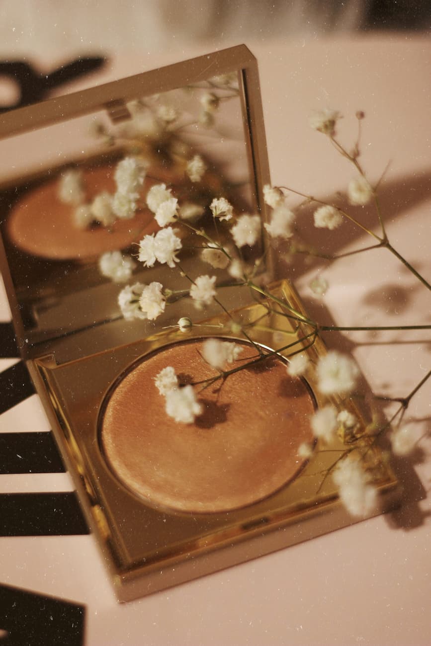 Golden compact highlighter with white dried flowers on a soft background. Elegant beauty product photography by Brenda Cazares, micropigmentation and tattoo expert in National City.