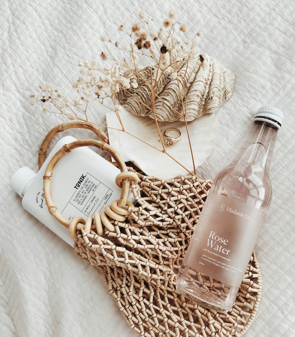 Flat lay of Madame Dry Rose Water, facial toner, and woven bag with natural textures. Beauty and skincare aesthetic by Brenda Cazares, tattoo and micropigmentation artist in National City.